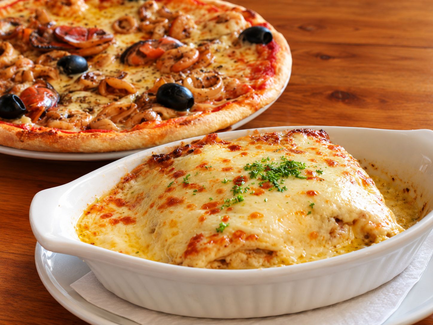 A bubbling dish of lasagna beside a seafood pizza with black olives